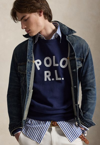 Jeune homme portant une veste en denim bleu superposée, un sweat-shirt bleu marine avec le texte « POLO R.L. », une chemise rayée, un pantalon blanc et une ceinture tressée marron.