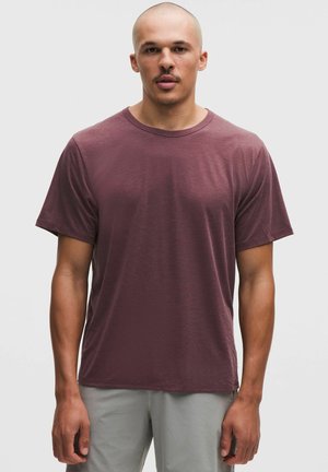 Uomo con testa rasata che indossa una maglietta a maniche corte color amaranto e pantaloni grigio chiaro, in piedi contro uno sfondo chiaro e uniforme.