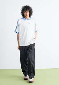 Polo shirt desportivo branco com detalhes em azul claro, combinado com calças de treino pretas. Apresenta o logótipo da Nike, corte solto e textura suave.
