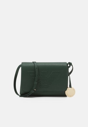 Sac bandoulière en cuir texturé vert foncé avec logo aigle en relief et étiquette ronde en or suspendue sur le côté.