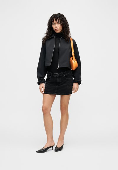Veste noire raccourcie avec zip, col roulé noir, jupe en denim noire et mules à talons noires. Sac mini orange porté à l'épaule.
