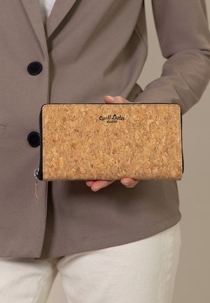 Korkpunge med en struktureret overflade, udstyret med en sort lynlås og et trykt logo. Holdes i hånden mod en neutral baggrund.