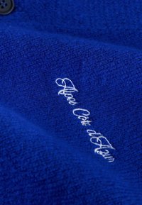 Blauwe fleece stof met een zachte textuur, voorzien van witte geborduurde tekst met de woorden "Alaïa Côte d'Azur." Zwarte knoopdetail zichtbaar.