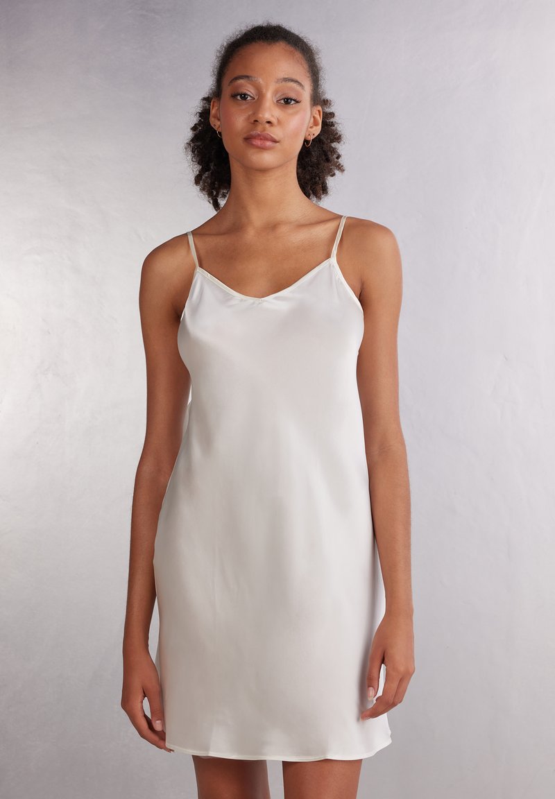 Vestido de satén en beige claro con finas tirantes tipo espagueti, escote en forma de v y una textura suave y fluida; hasta la rodilla con un ajuste ligeramente suelto.