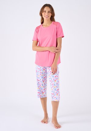 Vrouw staat blootsvoets, draagt een roze top met korte mouwen en kanten schouders en witte cropped broek met kleurrijke bladmotieven.