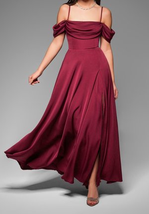 Femme portant une robe de soirée bordeaux fluide, épaules dénudées, avec une fente haute à la cuisse et des talons argentés, debout devant un fond gris uni.