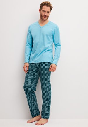 Mann trägt hellblaues langärmliges V-Ausschnitt-Oberteil und dunkelblaue gemusterte Pyjamahose, steht barfuß vor einer weißen Wand.