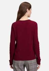 Kvinna med långt brunt hår, iklädd en bordeauxfärgad tröja och grå rutiga byxor, som står vänd bort från kameran mot en neutral bakgrund.