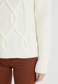Ivory sweter z dzianiny z wzorem w warkocze, z prążkowanymi mankietami i dołem, w połączeniu z brązowymi spodniami. Teksturowana włóczka i luźny krój zapewniają komfort.