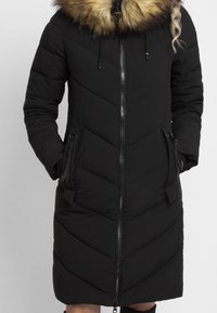 Abrigo de invierno negro largo y acolchado con cremallera frontal, dos bolsillos con cremallera y capucha forrada de piel sintética marrón, llevado con las manos en los bolsillos.