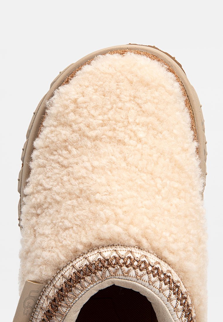 Zapatilla beige con un exterior de piel de oveja suave y costuras marrones. La suela es texturizada para mayor agarre, y el diseño presenta una punta redondeada.
