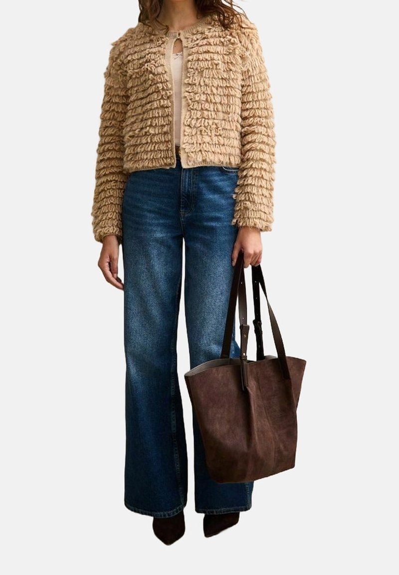 Femme portant une veste beige texturée et un jean bleu à jambes larges, tenant un grand sac cabas en suède marron à côté.