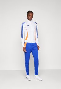 White sports jacket with orange accents and blue stripes, paired with blue pants featuring white stripes. Material appears smooth, athletic fit.