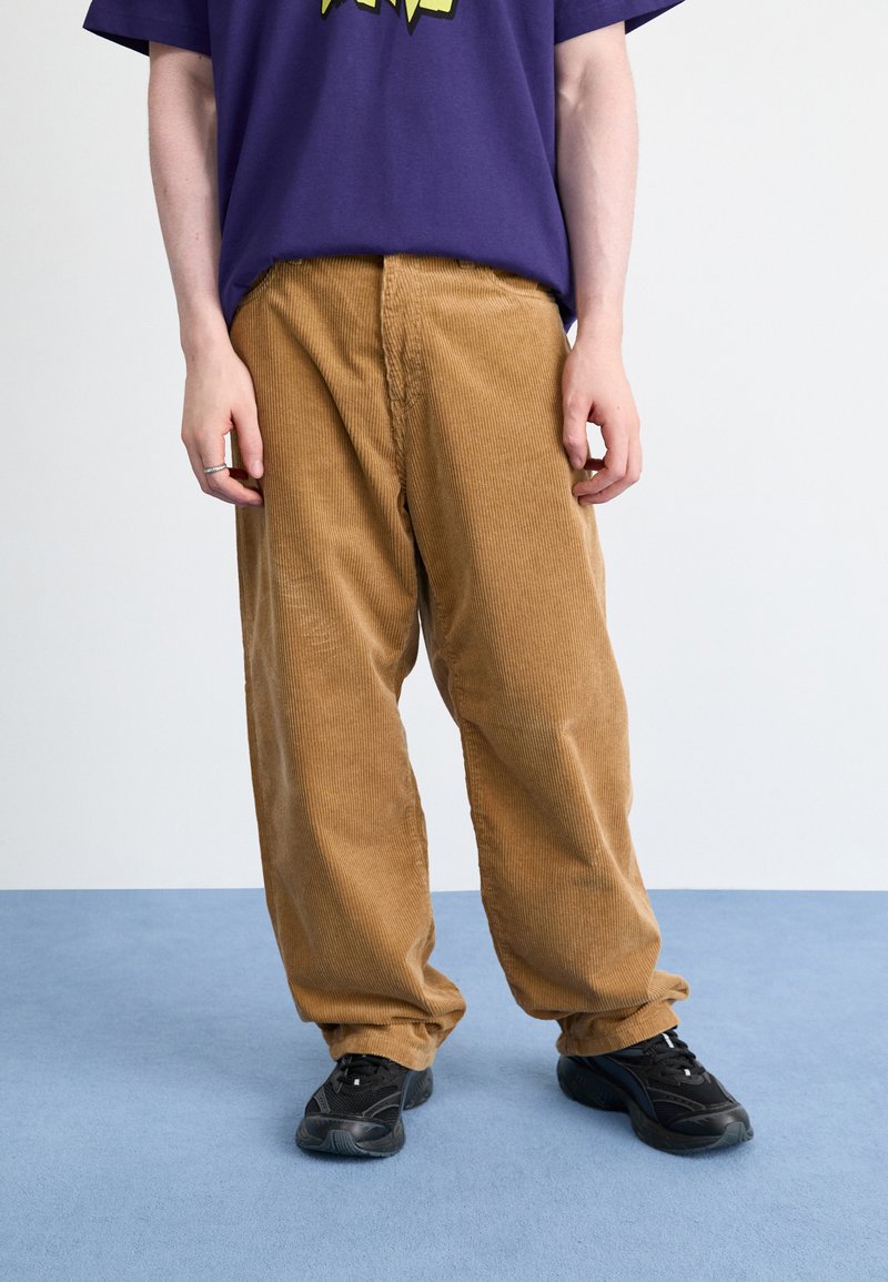 Brown corduroy pants with a relaxed fit, featuring vertical ridges. Worn with a purple graphic t-shirt and black athletic shoes.