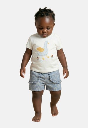 Tout-petit aux cheveux tressés portant une chemise blanche avec un motif de canard et un short à volants bleu fleuri, marchant pieds nus sur un fond uni.