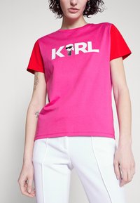 Růžové a červené tričko krátký rukáv se bílým textem "KRL" a grafickým potiskem. Měkká tkanina, uvolněný střih, lem mírně zakřivený.
