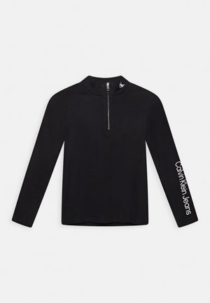Haut noir à manches longues avec une courte fermeture éclair devant, petit logo CK sur le col, et texte "Calvin Klein Jeans" sur la manche droite.