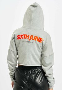 Kuttet grå hettegenser med en stor oransje "SIXTH JUNE" logo på ryggen. Har hette og en avslappet passform. Brukt med blanke svarte bukser.