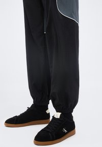 Άτομο που φοράει μαύρο παντελόνι τζόγκινγκ και μαύρα αθλητικά παπούτσια με καφέ σόλες και λευκό λογότυπο CK στο πλάι και στη γλώσσα.