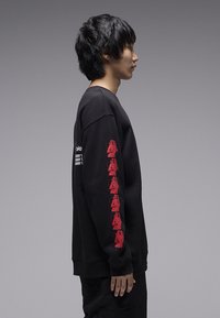 Μαύρο φούτερ με μακριά μανίκια, με κόκκινα γραφικά τυπωμένα κατά μήκος του αριστερού μανικιού. Μαλακή υφή, λαιμόκοψη με στρογγυλή λαιμόκοψη, άνετη εφαρμογή.