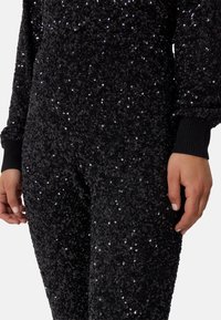 Svart paljett jumpsuit med en texturerad yta, ribbade manschetter och figurnära design. Närbild som betonar de glänsande, reflekterande paljetterna.