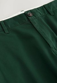 Pantalon vert foncé avec fermeture par bouton, passants pour ceinture et une ceinture intérieure blanche, montré en gros plan.