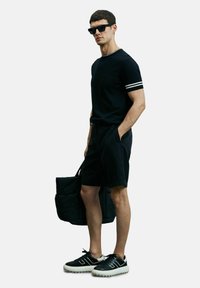 Zwarte T-shirt met witte dubbele streep op de mouwen, zwarte shorts, zwarte sneakers met witte accenten en een zwarte totebag.