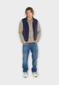 Gilet marine par-dessus une chemise gris clair, associé à un jean bleu. Les détails notables comprennent une fermeture éclair et des baskets avec un motif design.