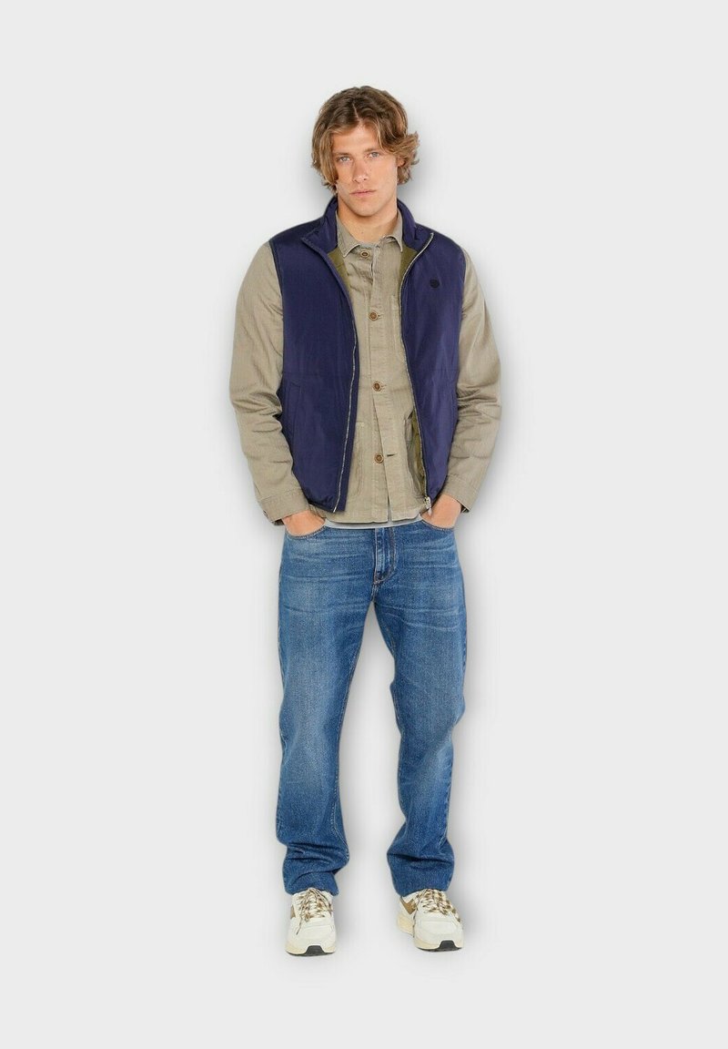 Gilet marine par-dessus une chemise gris clair, associé à un jean bleu. Les détails notables comprennent une fermeture éclair et des baskets avec un motif design.