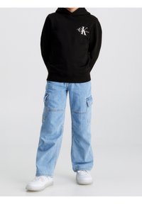 Felpa con cappuccio nera e logo bianco, abbinata a pantaloni cargo blu chiaro con tasche laterali e sneakers bianche.