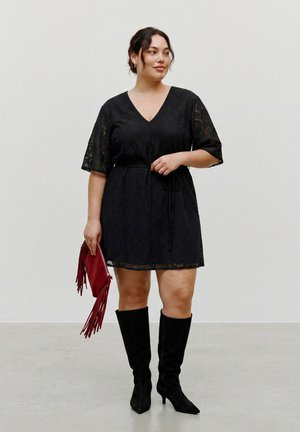 Femme portant une courte robe noire en dentelle avec des manches aux coudes, des bottes noires montantes jusqu'aux genoux, tenant une pochette rouge à franges.