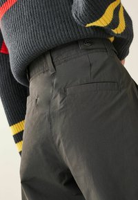 Donkergrijze broek van glad, lichtgewicht materiaal. Bevat een knoopsluiting, achterzakken en subtiele stikdetails.