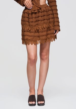 Mini-jupe marron texturée avec bordure en dentelle, associée à des sandales mule noires à bouts ouverts sur un mannequin debout devant un fond uni.