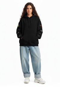 Felpa nera con tasca frontale, dettagli grafici sulle maniche e vestibilità morbida. Abbinata a jeans baggy azzurri e sneakers bianche.