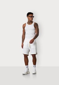 Weißes Tank-Top, weiße Shorts mit Struktur und Kordelzug sowie weiße Sneakers. Zu den Accessoires gehören dunkle Sonnenbrillen und gemusterte Socken.