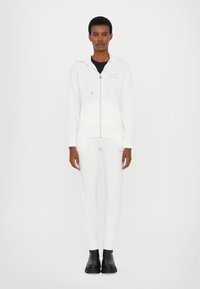 Biała bluza z kapturem na zamek błyskawiczny z troczkami i logo, zestawiona z białymi slim-fit spodniami. Model nosi czarne botki za kostkę, stojąc na jednolitym tle.
