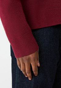 Burgundowy sweter dzianinowy z prążkowanymi mankietami, pokazujący teksturowany materiał oraz zbliżenie ręki opierającej się na ciemnym dżinsie.