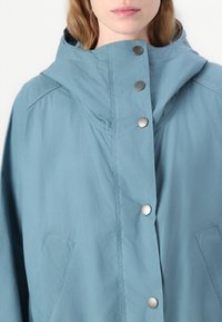 Blue lightweight jacket with a hood, snap buttons, two side pockets, and a smooth texture. Features stitching along the shoulders.