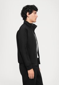Jeune homme aux cheveux bouclés portant une veste zippée noire et un pantalon noir, se tenant de profil contre un fond blanc uni.