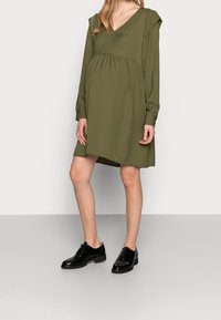 Olivi vihreä mekko, jossa on V-pääntie ja pitkät hihat, vyötärössä on kootut yksityiskohdat ja olkapäissä korostuksia. Päällä mustat kiiltävät nahkakengät.