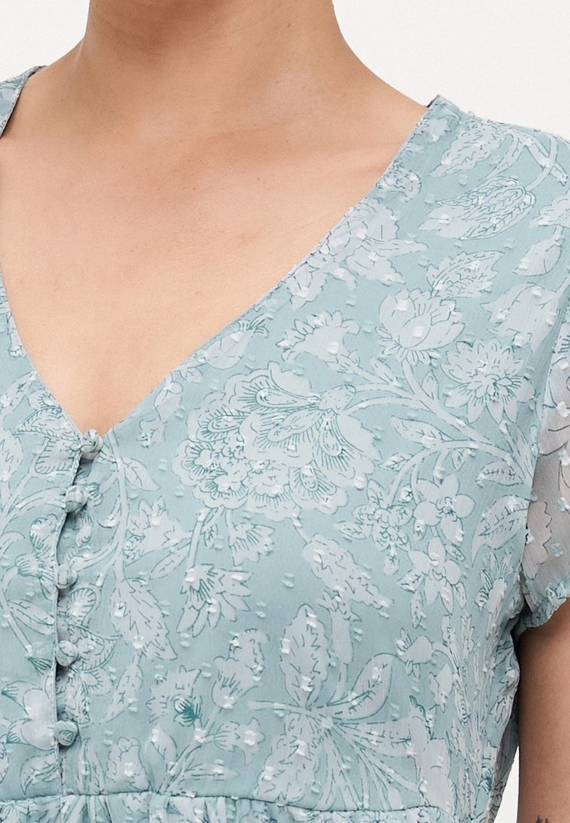 Robe fleurie bleu clair en tissu texturé avec de petits boutons sur le devant, portée par une personne avec le cou et le haut de la poitrine dégagés.