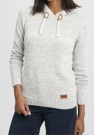 Woman wearing light gray knit hooded sweater with wooden toggle details and ribbed cuffs, paired with dark blue corduroy pants.