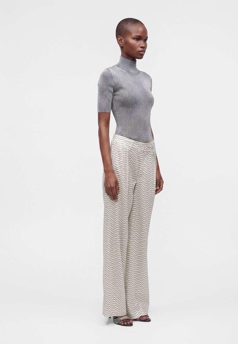 Femme vue de profil portant un haut gris à manches courtes et col roulé, un pantalon large à motifs et des sandales à brides sur fond blanc.