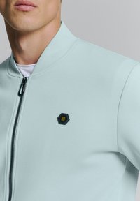 Lichtblauwe ritssluitingjas met een textuurafwerking, met een zwart hexagonaal logo op de borst en een ontwerp met hoge kraag.