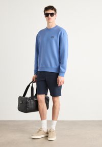 Light blue sweatshirt with ribbed cuffs, navy shorts, beige sneakers, and white socks. Black duffel bag with straps and silver accent.