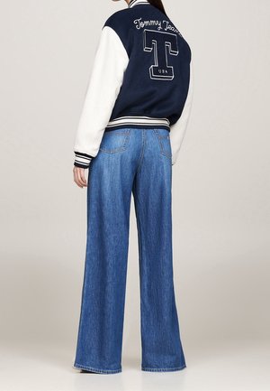 Vrouw draagt een navyblauwe en witte Tommy Jeans varsityjas en wijde blauwe spijkerbroek, staand met haar rug naar de camera tegen een effen achtergrond.