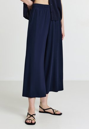 Pantalon large bleu marine en tissu lisse, doté d'une ceinture élastique et de poches latérales, assorti à des sandales noires minimalistes.