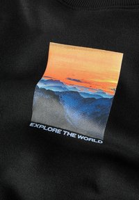 Crni dukserica s pravokutnim grafičkim prikazom zalaska sunca iznad planina, s nijansama narančaste, plave i sive boje. Natpis glasi "ISTRAŽUJ SVIJET."