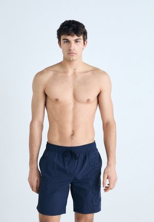 Navy blue swim shorts with an elastic waistband and drawstring, featuring a prominent embossed logo detail on the left leg.