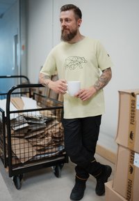 Light green t-shirt features a geometric bear design. Black cargo pants and sturdy black shoes with orange accents. Background has a cart of materials.